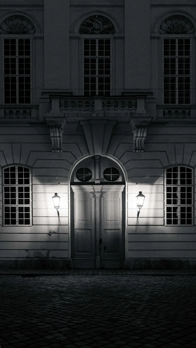 Baroque palace entrance at night — symmetrical twin lanterns, cobblestone, black and white by Peter Scholz