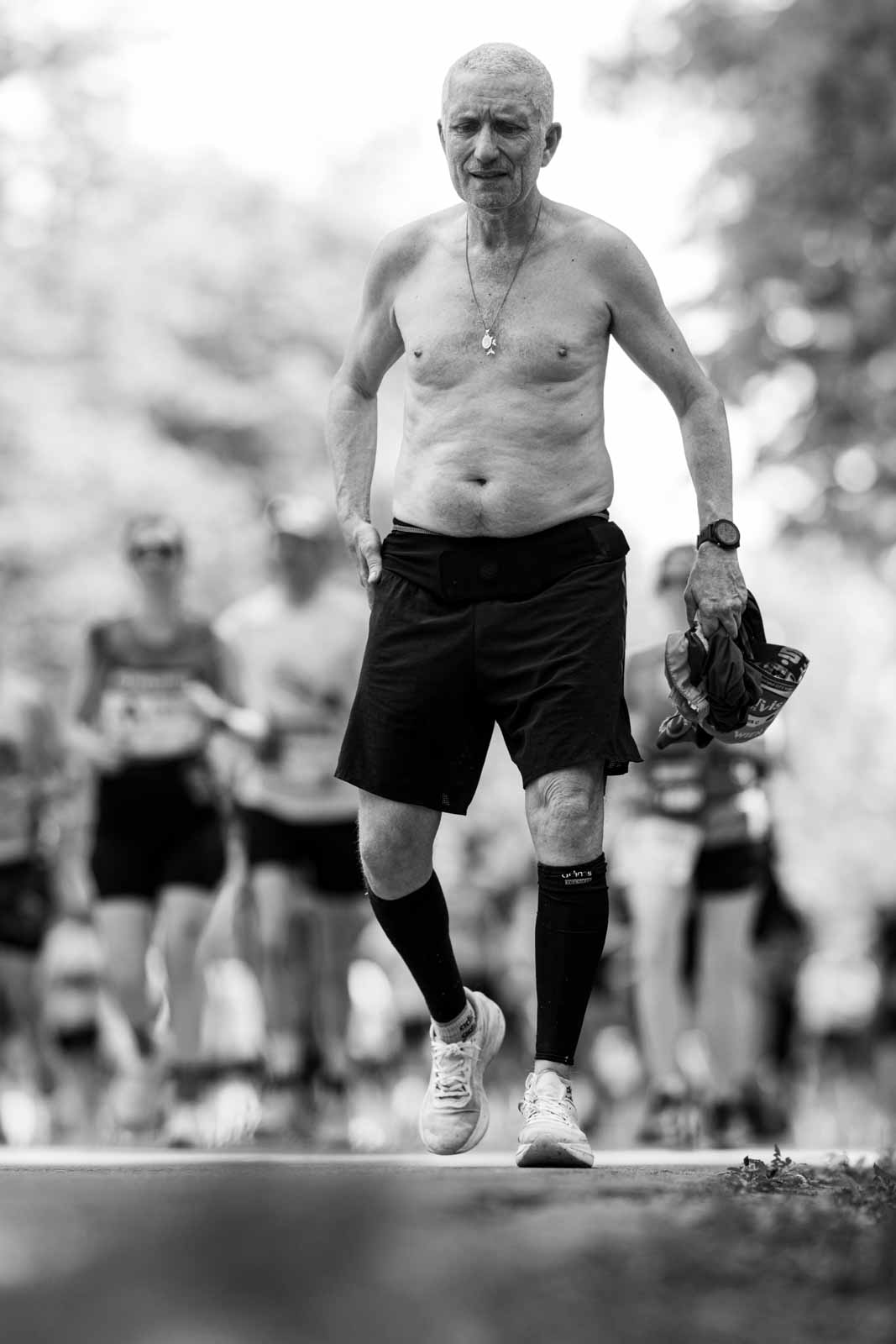 Shirtless senior marathon runner with compression sock and necklace walking through the Vienna Prater — black and white fine art photography by Peter Scholz