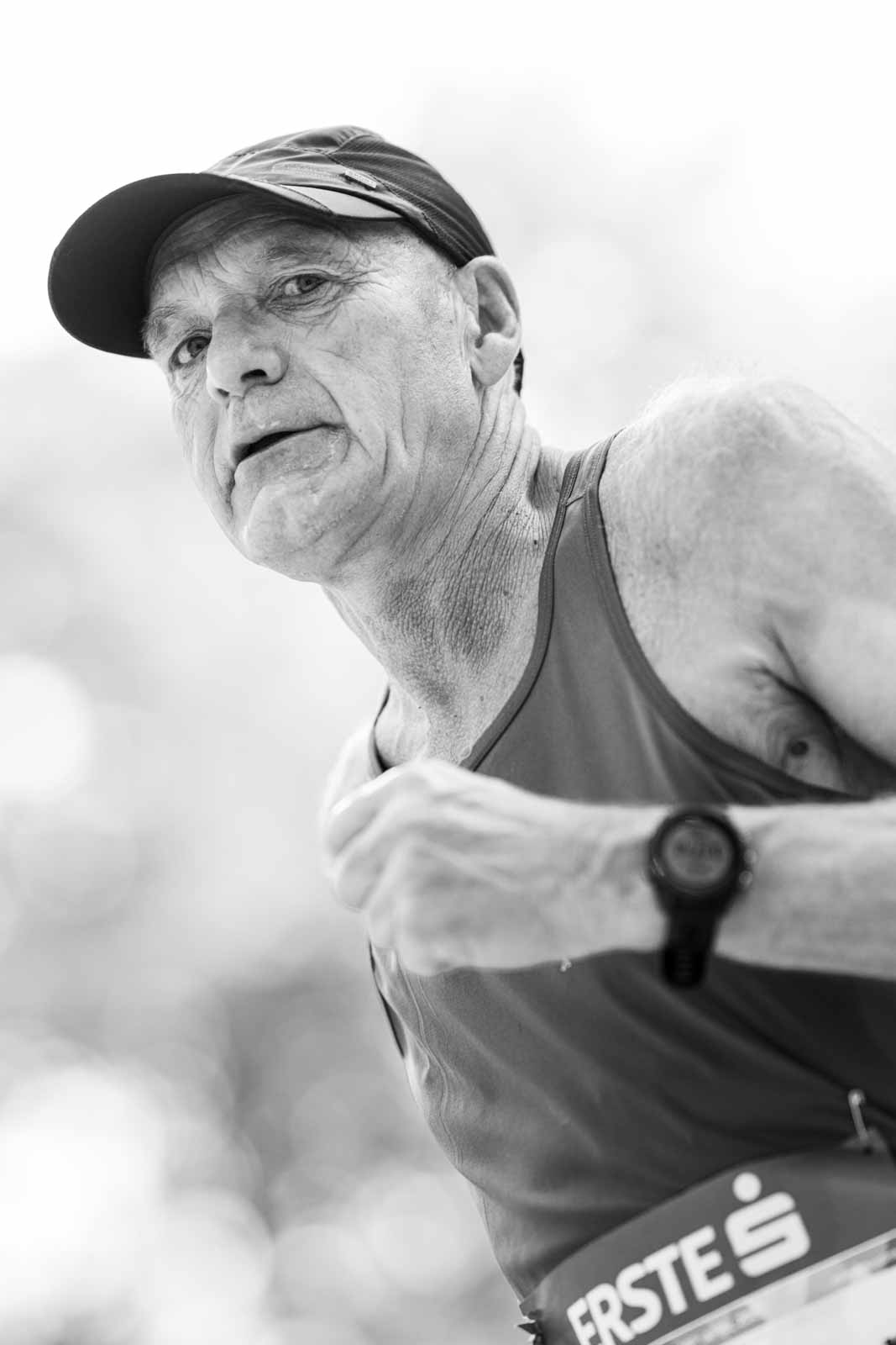 Senior marathon runner with cap in full effort at the Vienna City Marathon — black and white low-angle portrait photography by Peter Scholz